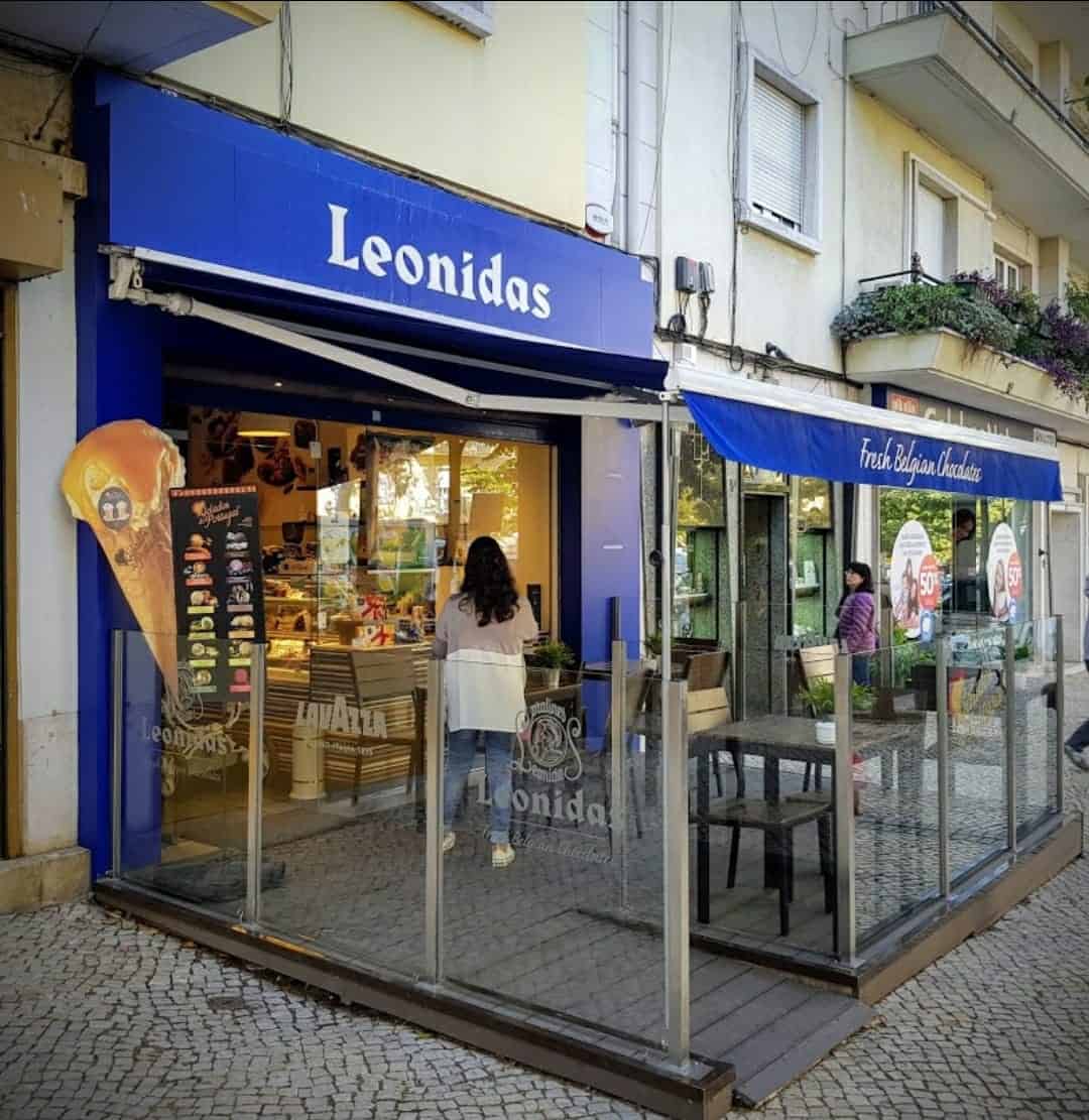 Loja física da Leonidas/ ponto de venda em Portugal em Lisboa Alvalade. Esplanada envidraçada em frente à montra de vidro com letreiros em azul com o logótipo da Leonidas.